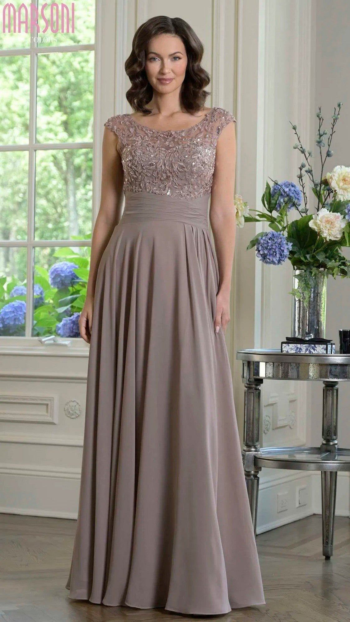 A woman stands indoors near a window and floral arrangements, wearing the Marsoni M335 long formal dress in taupe—a floor-length chiffon A-line gown with a lace bodice and cap sleeves.